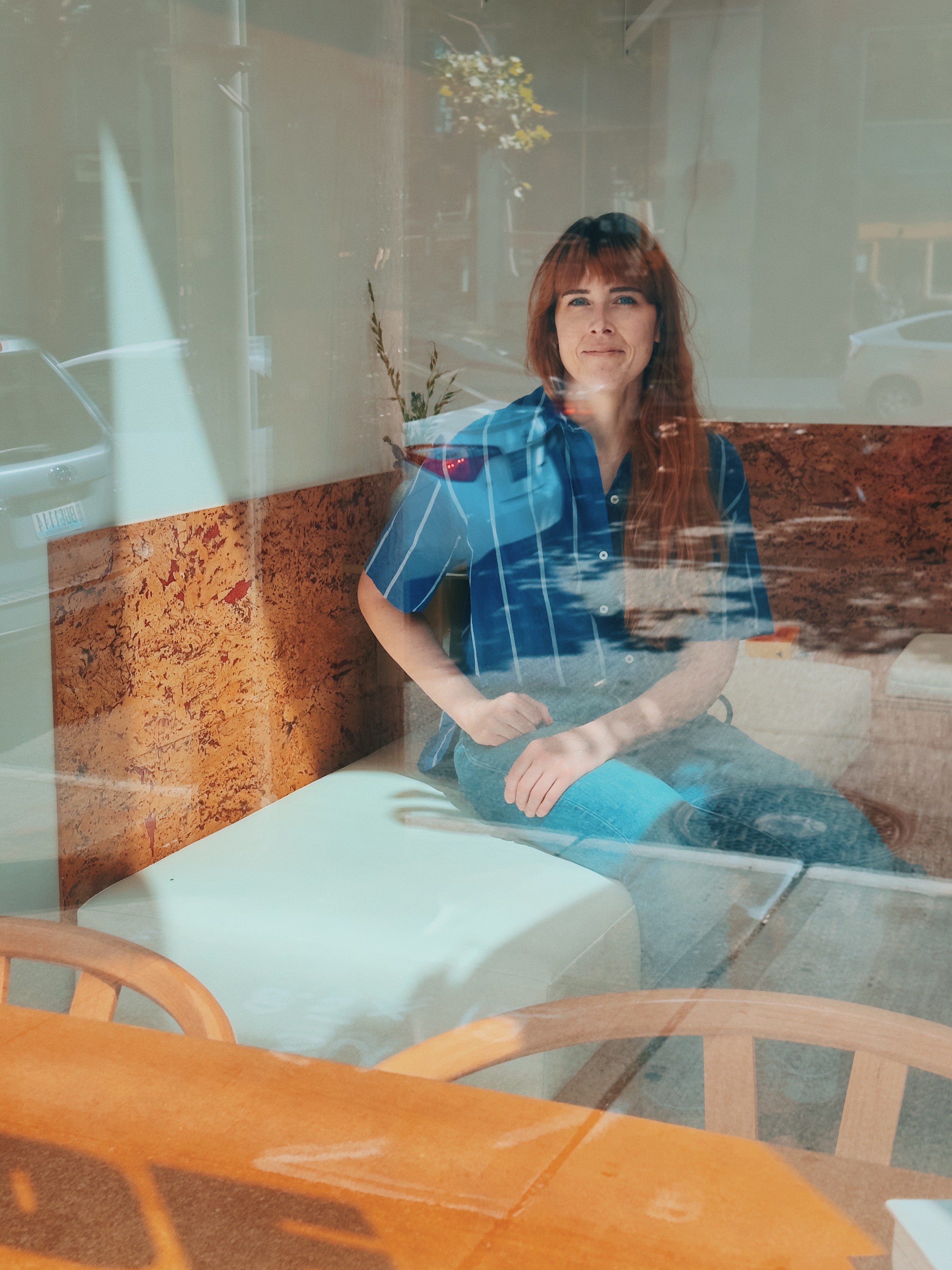 Leslie, a white woman with long, red hair in a blue-striped collared shirt sits inside a coffee shop and is photographed through the window.